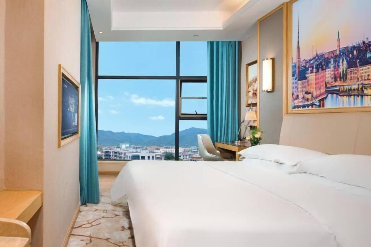 A hotel room with a large bed, a desk, a chair, a window with a view of a cityscape, and a painting on the wall.