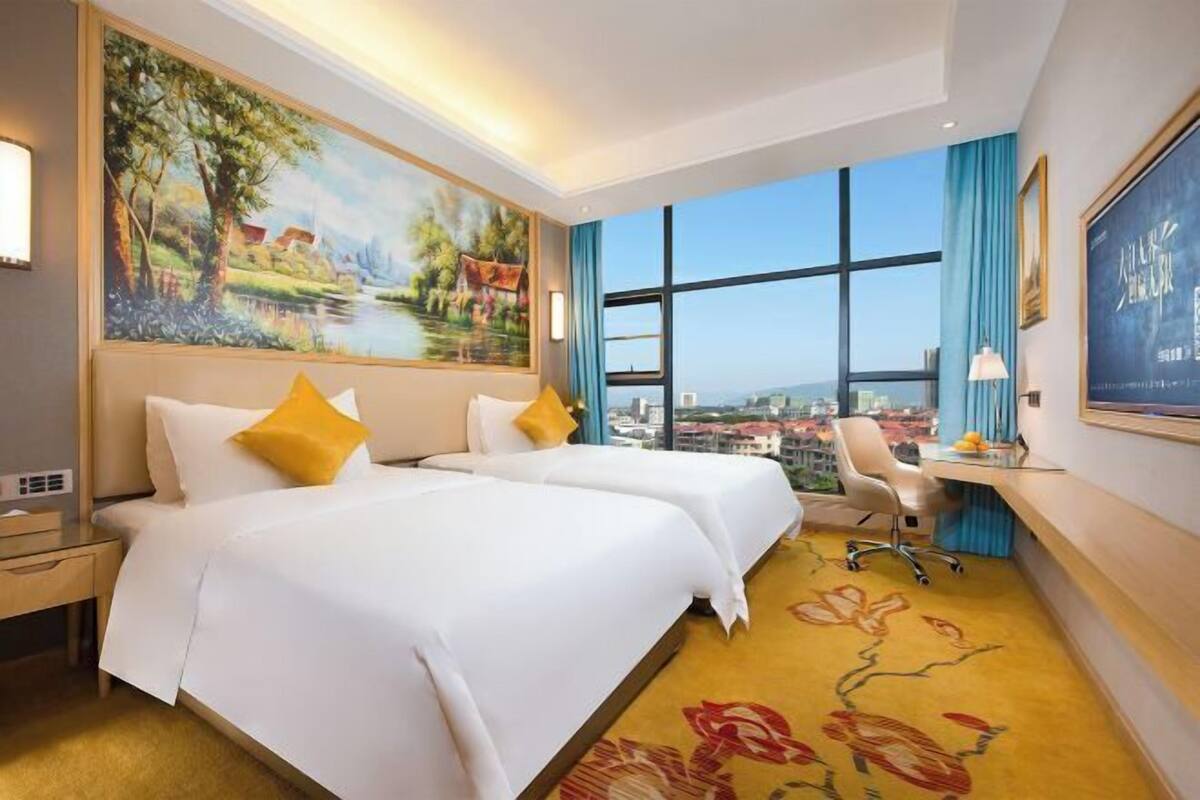 A hotel room with two beds, a desk, a chair, a large window with a city view, and a painting on the wall.