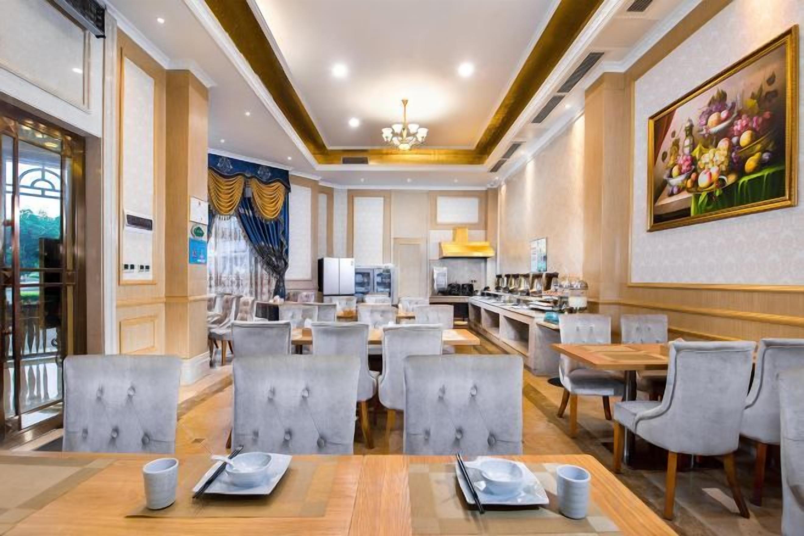 A hotel dining area with multiple tables and chairs, a kitchen in the background, and a painting on the wall.