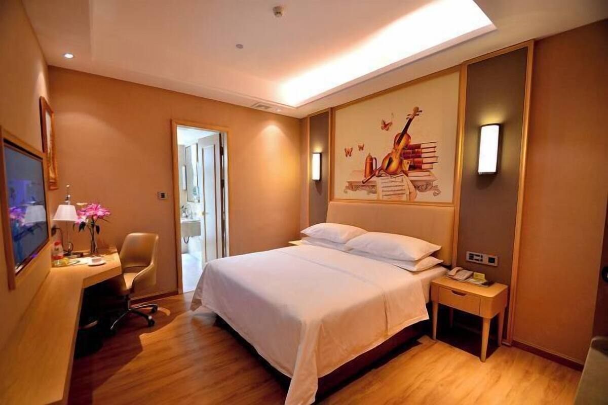 A hotel room with a bed, bedside table, desk with a lamp, chair, and a painting on the wall.