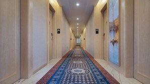 Un couloir d’hôtel avec un tapis à motifs, des portes des deux côtés et un passage menant à un ascenseur.