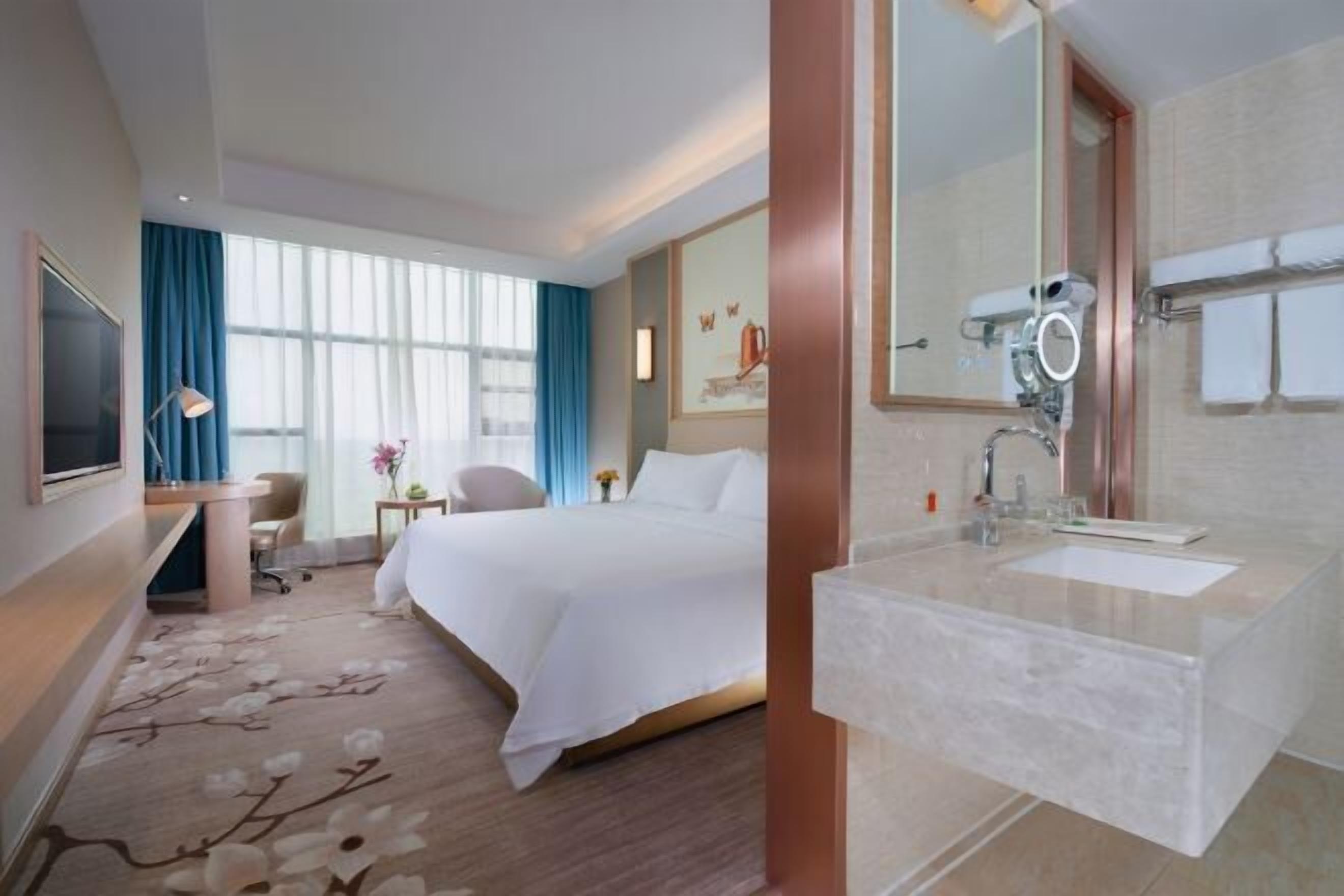 A hotel room with a bed, a desk with a chair, a large window with curtains, a bathroom with a sink and mirror, and a wall-mounted TV.