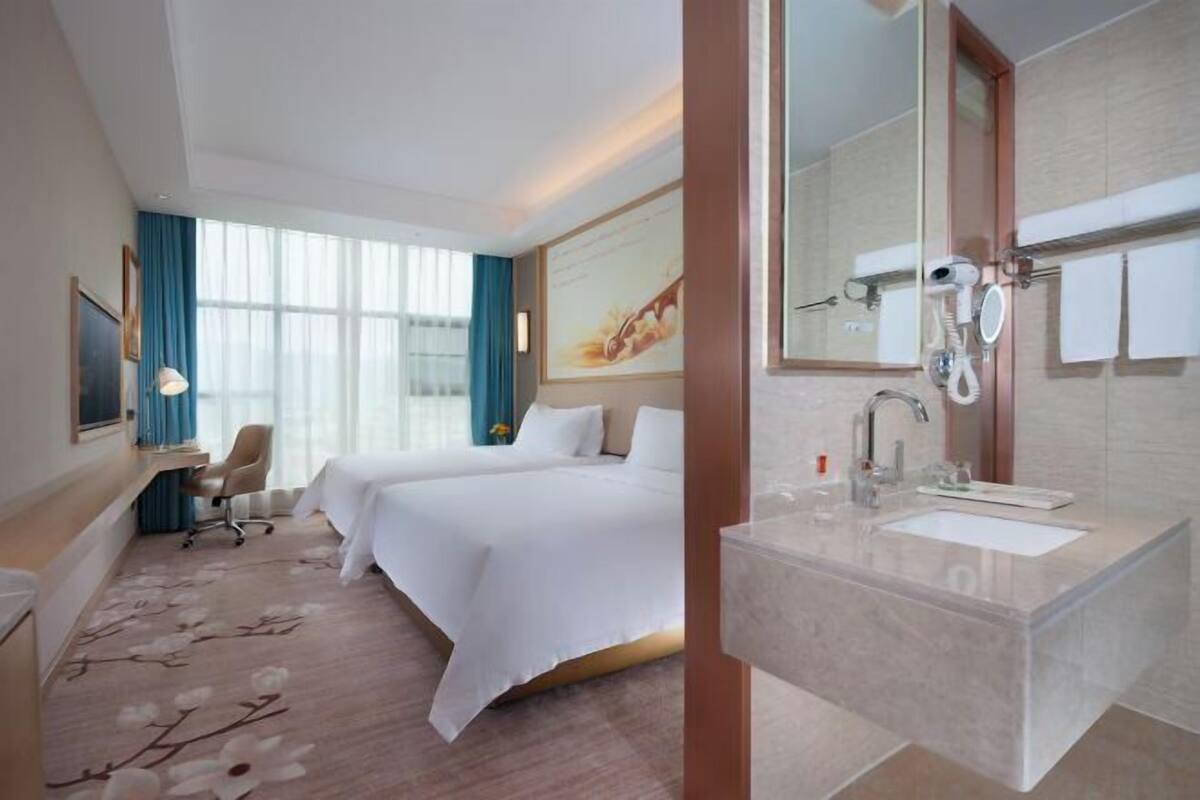 A hotel room with a large bed, a desk, a chair, a bathroom with a sink and mirror, and a window with curtains.