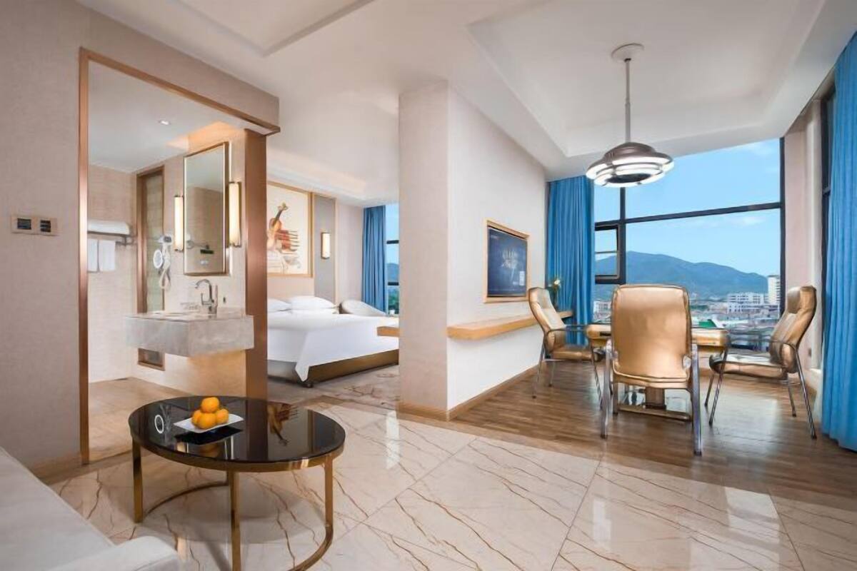 A hotel room with a bed, a sitting area with a table and chairs, a large window with a view, and a bathroom with a mirror and sink.
