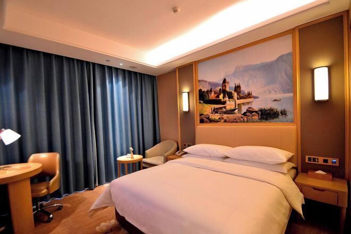 A hotel room with a large bed, a desk, a chair, and a painting of a scenic landscape on the wall.