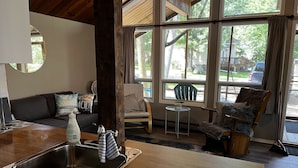 Un accogliente soggiorno con soffitto in legno, un divano con cuscini, una poltrona, un tavolino e un'ampia finestra con vista sull'esterno.