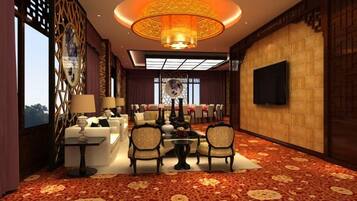 A hotel lobby with a large chandelier, a seating area with tables and chairs, a television mounted on the wall, and a patterned carpet.