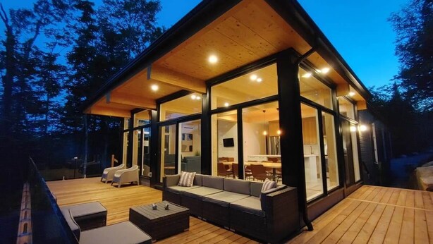 Un espace de vie extérieur moderne, doté d’une terrasse en bois, de sièges intégrés et de grandes portes vitrées donnant sur une salle à manger intérieure.