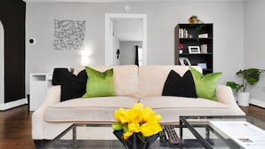 A modern living room with a beige sofa, green and black cushions, a glass coffee table with a vase of yellow flowers, a black bookshelf, and a white side table.