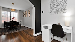 A dining area with a wooden table and chairs, a black chair, a white desk with a black chair, a wall-mounted abstract art piece, and a hanging light fixture.