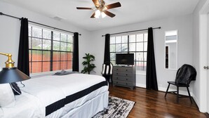 A bedroom with a bed, a ceiling fan, a TV, a dresser, a chair, and large windows with blinds.