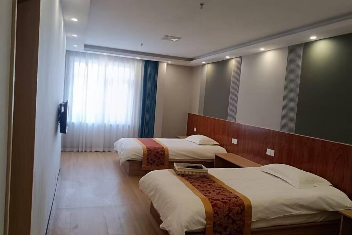 A hotel room with two beds, a wooden headboard, a bedside table, and a window with curtains.
