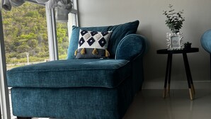 A teal sofa with a patterned pillow, a small side table with a plant, and a view of greenery outside. - AC, WiFi, cable & 24/7 security, Pool, beachfront, views of Kingston harbor. (Portmore)