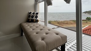 A modern room with a large window, a tufted bench, and a view of the sea. - AC, WiFi, cable & 24/7 security, Pool, beachfront, views of Kingston harbor. (Portmore)