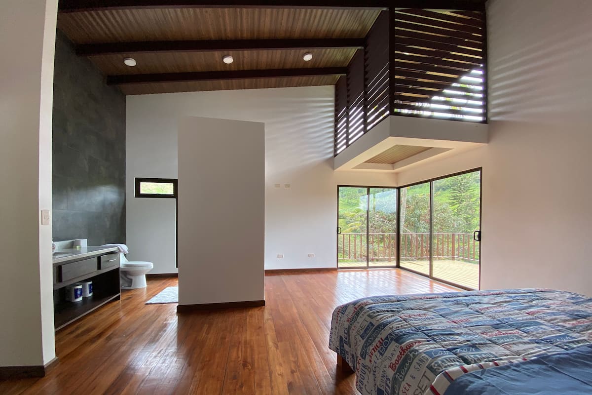 Une chambre moderne avec un grand lit, un plancher en bois, un banc intégré et un balcon donnant sur un paysage verdoyant.