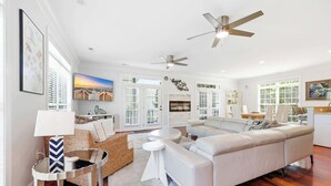 A spacious living room with a large white sofa, a coffee table, and a TV. There are ceiling fans, a fireplace, and large windows. - Sun and Games House w Swimming Pool Tons of Games Putting Green (North Myrtle Beach)