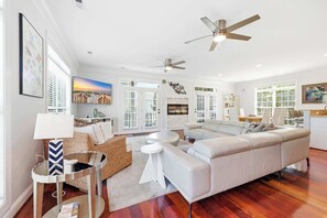 Un salon spacieux avec un grand canapé blanc, une table basse et un téléviseur. On y trouve également des ventilateurs de plafond, une cheminée et de grandes fenêtres.
