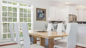 A modern kitchen with white cabinets, a stainless steel refrigerator, and a dining area with a wooden table and white chairs. - Sun and Games House w Swimming Pool Tons of Games Putting Green (North Myrtle Beach)