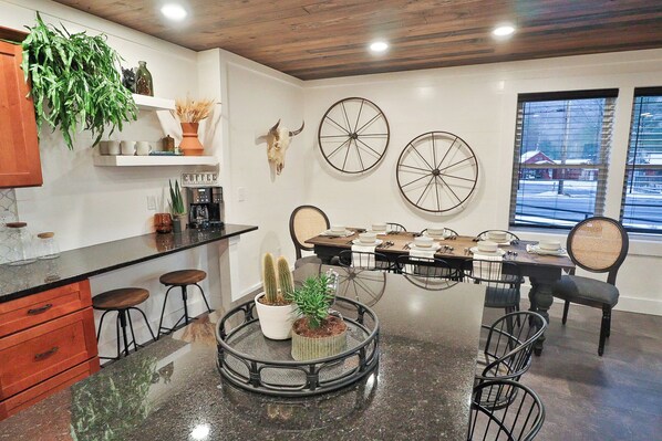 A modern kitchen with a central island, a dining area with a large wooden table, and a window with a view of a snowy landscape. - The 1849 Loft (Lake George)