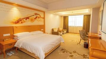 A hotel room with a large bed, a desk, a chair, and a sofa. There is a painting on the wall and a window with curtains.