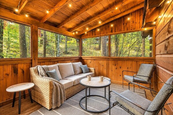 A wooden cabin interior with a sofa, two chairs, a small table, and large windows offering a view of the forest.