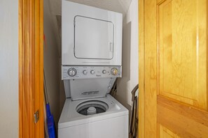 Eine kompakte Wasch- und Trocknerkombination in einem kleinen Hauswirtschaftsraum mit einer Holztür und einem blauen Wischmopp, der an der Wand lehnt.