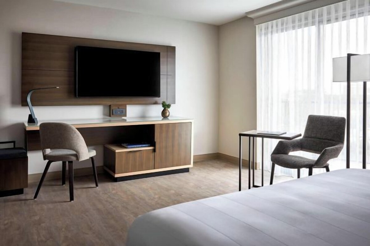 Ett modernt hotellrum med en stor säng, ett skrivbord med en stol, en tv som är monterad på väggen och ett fönster med tunna, genomskinliga gardiner.