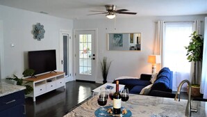 A modern living room with a TV, seating area, and a dining table with wine glasses and a bottle. - The Blue Bungalow by Crescent Lake Walk 2 Downtown K&Q Hot Tub Private Courtyard (St. Petersburg)