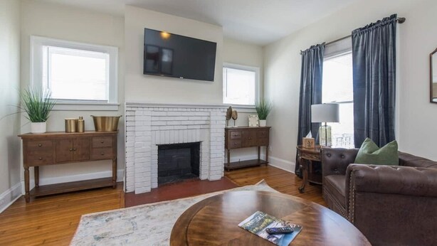 A living room with a fireplace, a flat-screen TV mounted above it, a brown leather sofa, and a coffee table with a magazine.
