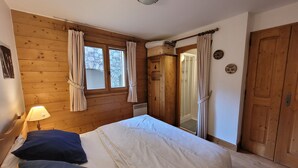 Une chambre avec un plancher en bois, un lit avec une literie blanche et bleue, une table de chevet, une lampe, une fenêtre avec des rideaux et une armoire.