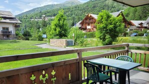 Une terrasse en bois avec une table et des chaises, d’où l’on peut admirer un paysage verdoyant, avec des maisons et des montagnes en arrière-plan.