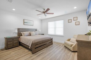 A bedroom with a bed, nightstands, a ceiling fan, a window with blinds, and a small seating area.