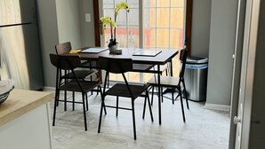 Un coin repas avec une table et des chaises, une fenêtre donnant sur une clôture en bois et une poubelle.