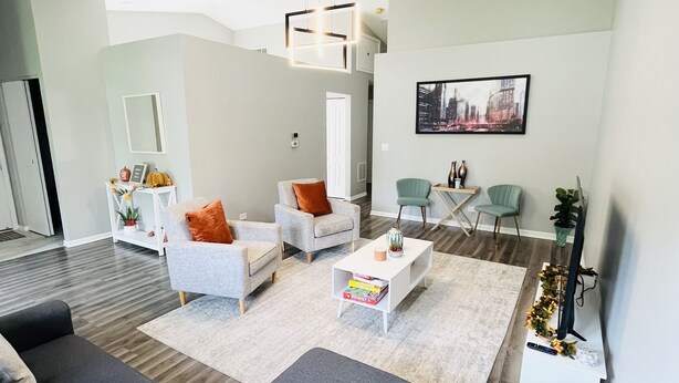 Un salon moderne avec un canapé gris, des fauteuils blancs et une table basse. On y trouve un téléviseur posé sur un meuble, une bibliothèque et un tableau accroché au mur.