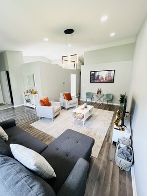 Una sala de estar moderna con un sofá gris, sillones blancos y una mesa de centro. Hay un televisor sobre un mueble bajo, una estantería y un cuadro en la pared.