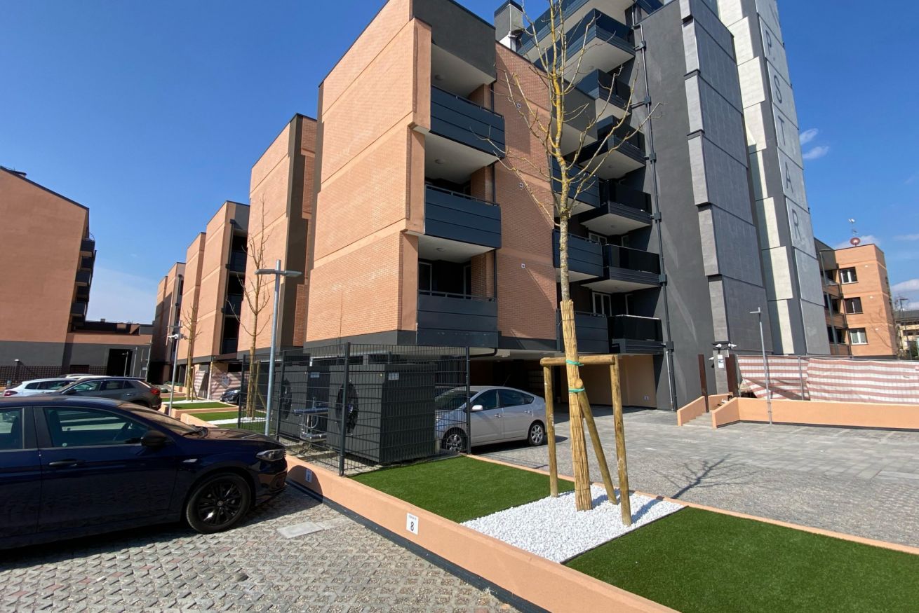 Un edifici d'apartaments de diverses plantes amb balcons, una zona d'aparcament i un arbre petit.