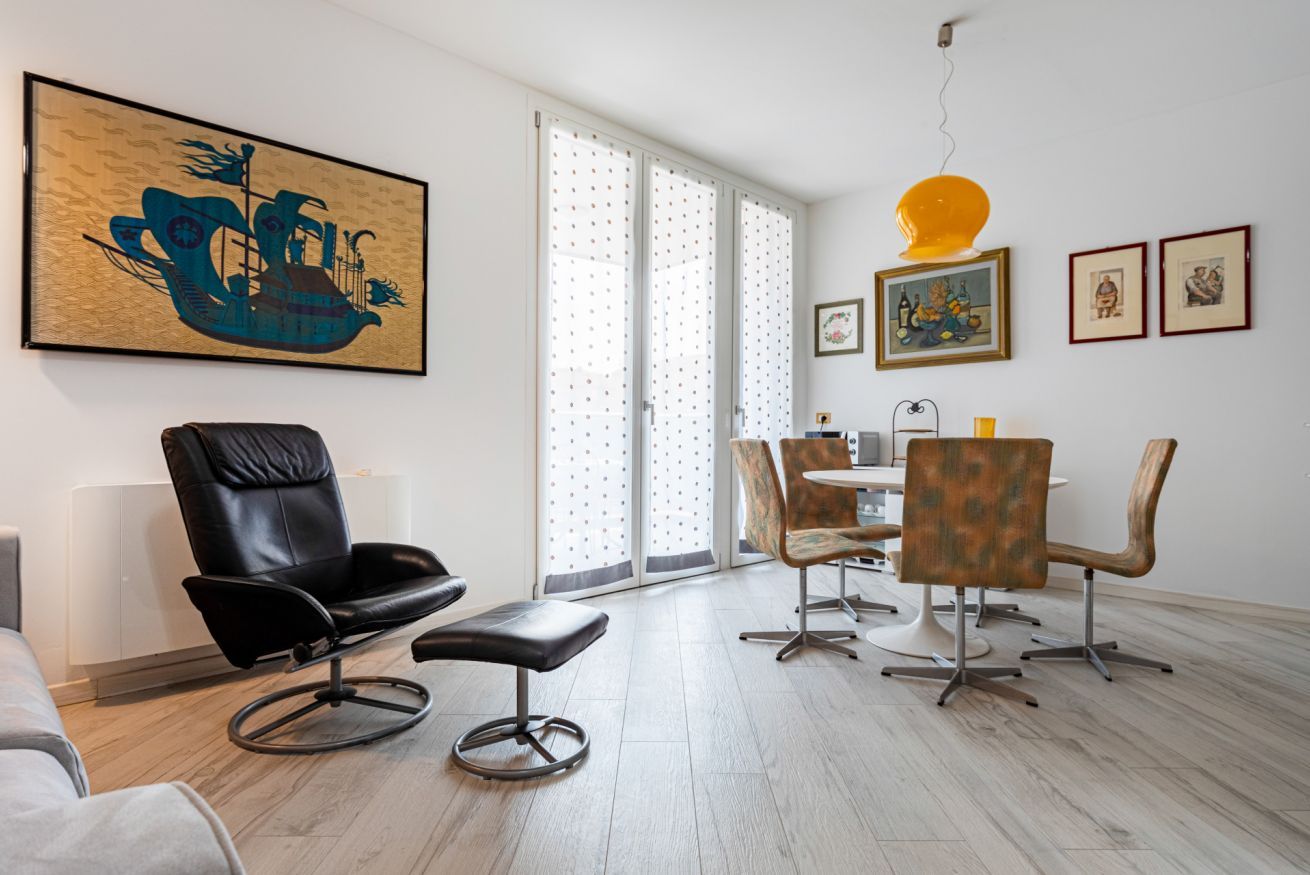 A modern living room with a black leather chair, a dining table with chairs, a large abstract painting, and a yellow pendant light.
