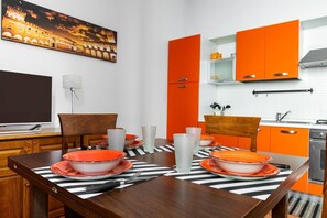 A modern kitchen with orange cabinets, a dining table set with orange plates and glasses, and a painting of a historical building on the wall.