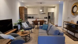 A modern living room with a sofa, chairs, a coffee table, and a TV. There is a dining area with a table and chairs. The kitchen is visible in the background with cabinets and appliances. - Cozy apartment, full equipped, in a gated community, close to places of interest (Sint Michiel)