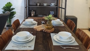 A dining area with a wooden table set for four, surrounded by chairs. A metal shelf unit with glassware and dishes is behind the table. - Cozy apartment, full equipped, in a gated community, close to places of interest (Sint Michiel)