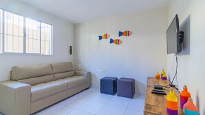 Un salon avec un canapé beige, deux poufs, une table basse en bois ornée de bouteilles colorées, un téléviseur à écran plat et des décorations murales représentant des poissons.