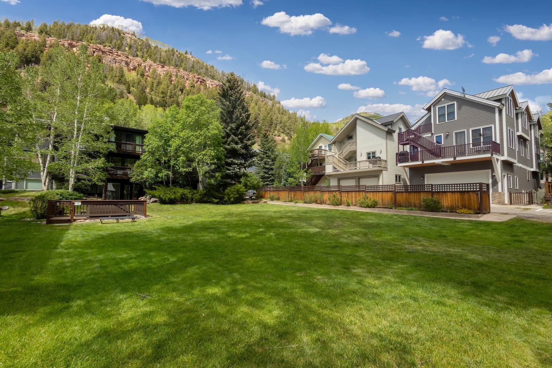A residential area with a large house featuring a balcony, a well-maintained lawn, and a forested hillside in the background.