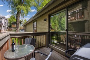 Una terraza cubierta al aire libre con una mesa y sillas de vidrio, una botella de vino y vistas a una zona residencial con casas y árboles.