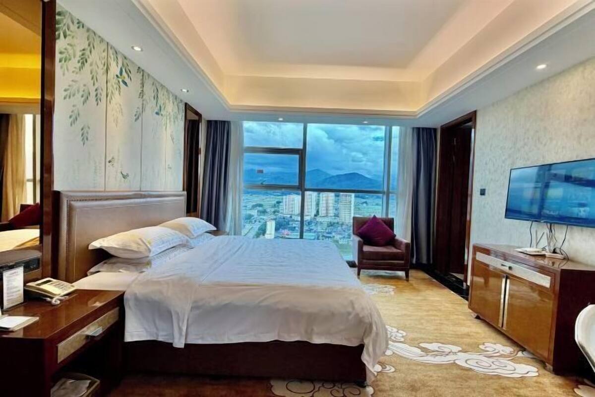A hotel room with a large bed, a desk with a telephone, a TV, and a view of the city through large windows.