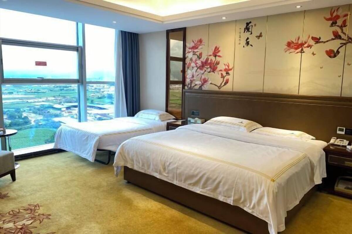 A hotel room with two beds, a large window offering a view of the countryside, and a traditional-style wall with floral artwork.