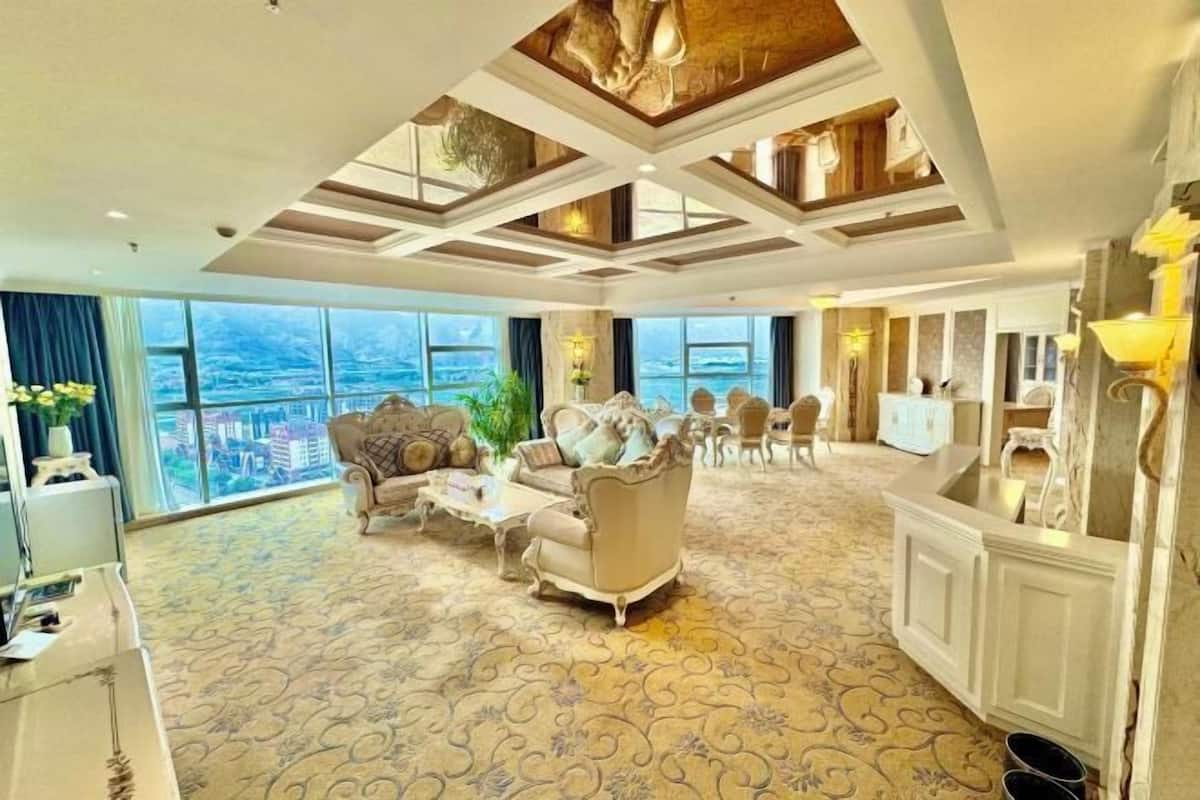A spacious hotel room with a large window offering a city view, a classic armchair, a sofa, and a dining area with a chandelier.
