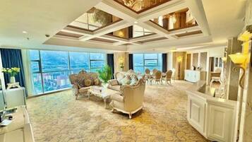 A spacious hotel room with a large window offering a city view, a classic armchair, a sofa, and a dining area with a chandelier.