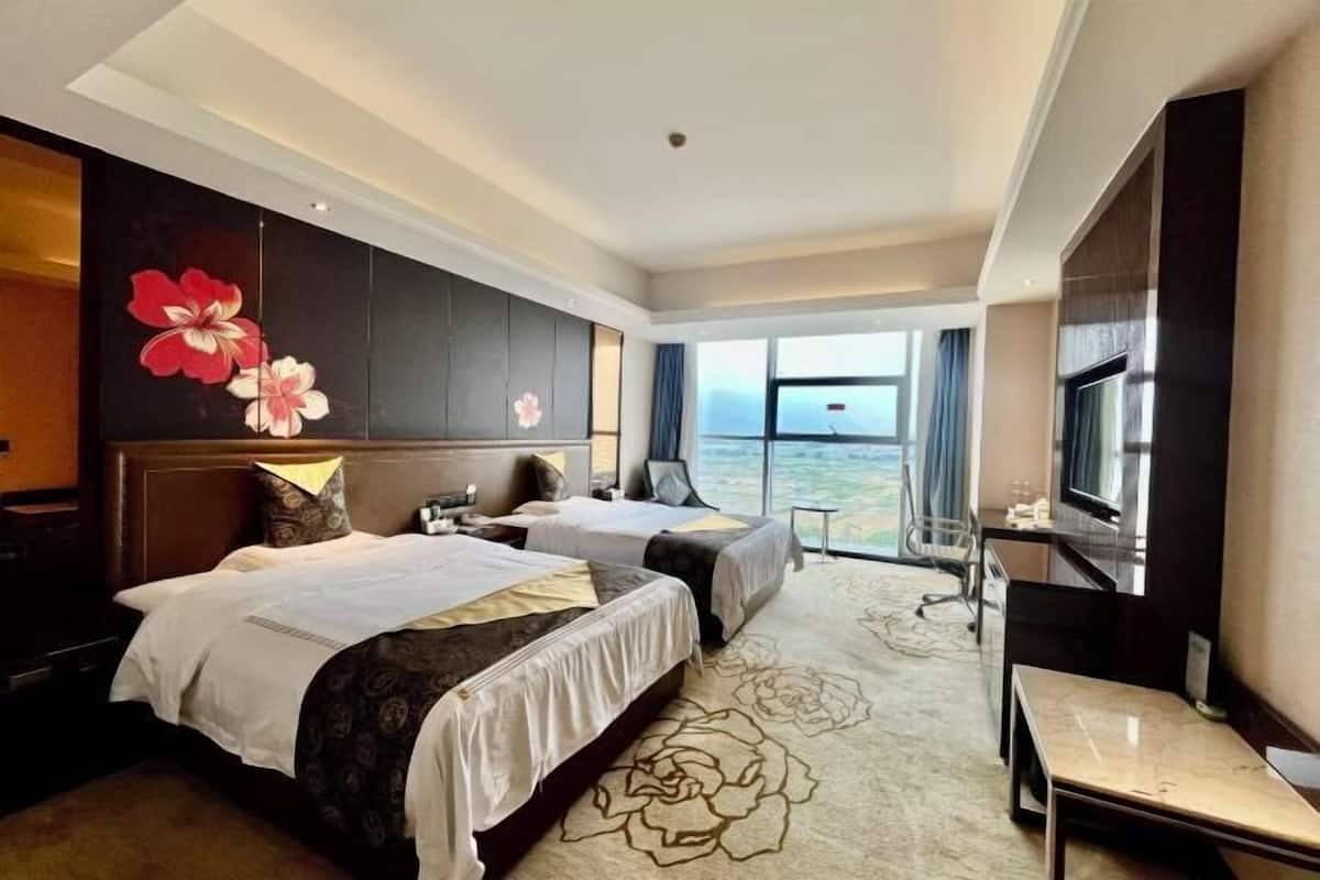 A hotel room with two beds, a desk, a chair, a large window with a view, and a decorative wall panel.