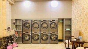 A hotel laundry room with multiple washing machines, a small table with a chair, and a trash bin. - Hongbo Hotel (Gengma)
