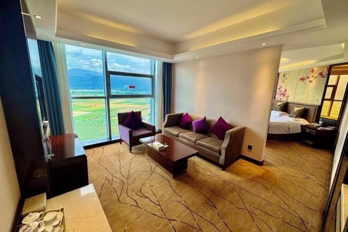 A modern hotel room with a large window offering a view of the outdoors, a sofa with purple cushions, a small coffee table, and a bed with a nightstand.